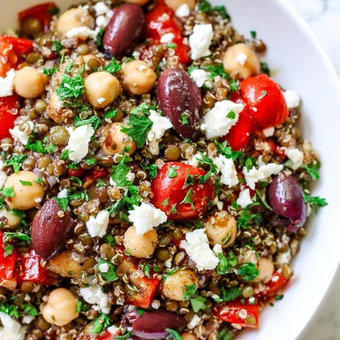 A close-up of Greek Power Salad in a white bowl, showing lentils, quinoa, and chickpeas mixed with colorful diced vegetables, topped with feta and olives.
